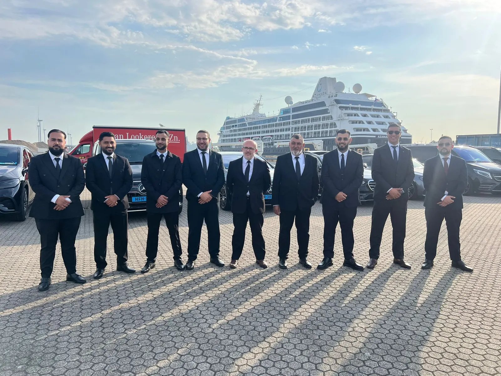 Nine chauffeurs in dark suits and ties standing in a row in front of Mercedes cars and a cruise ship
